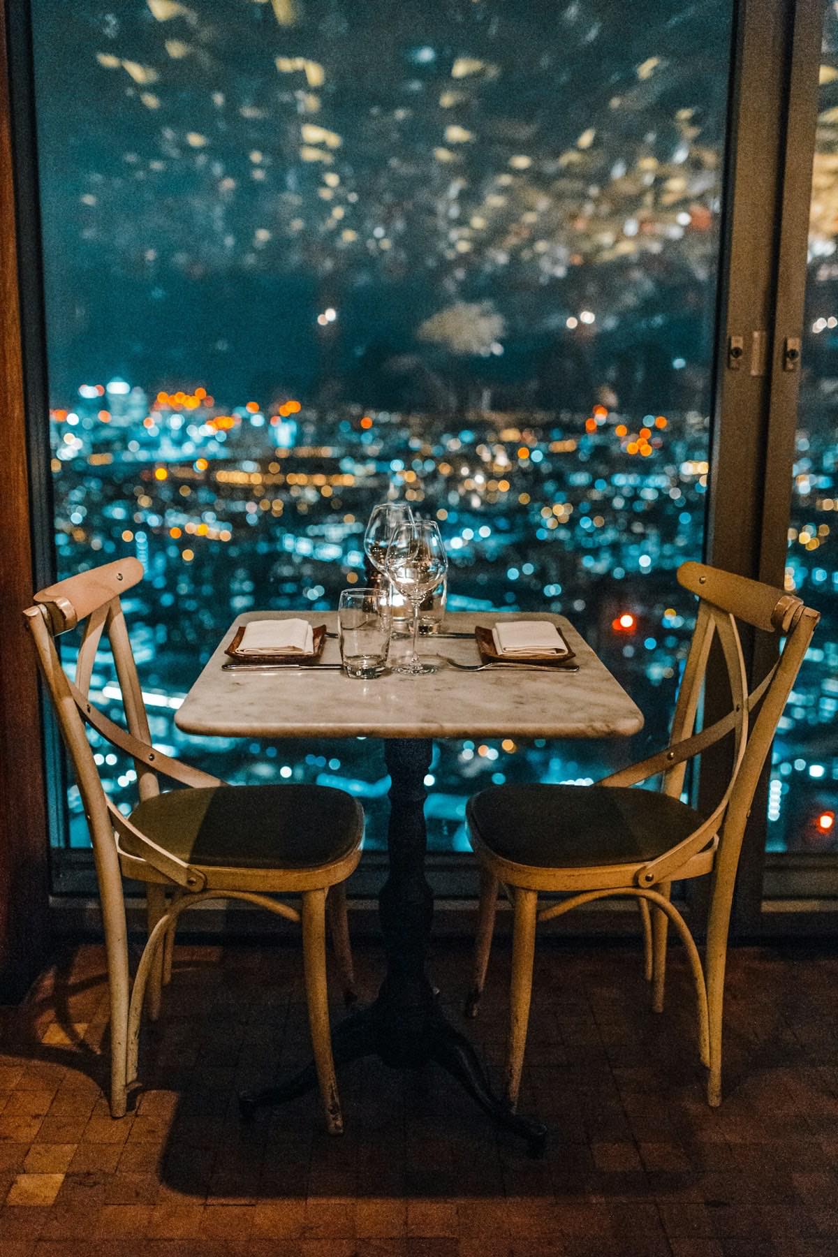 A table set for two by a window, above a city at night